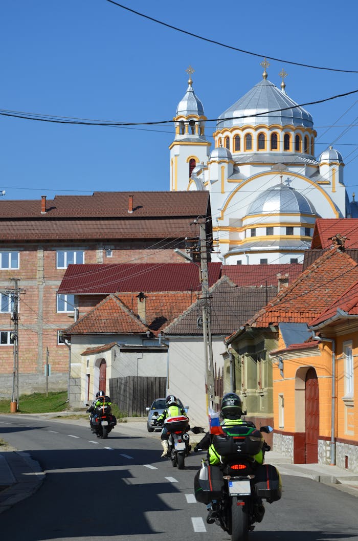 Motorcyclists journey through Romanian village street with church backdrop.